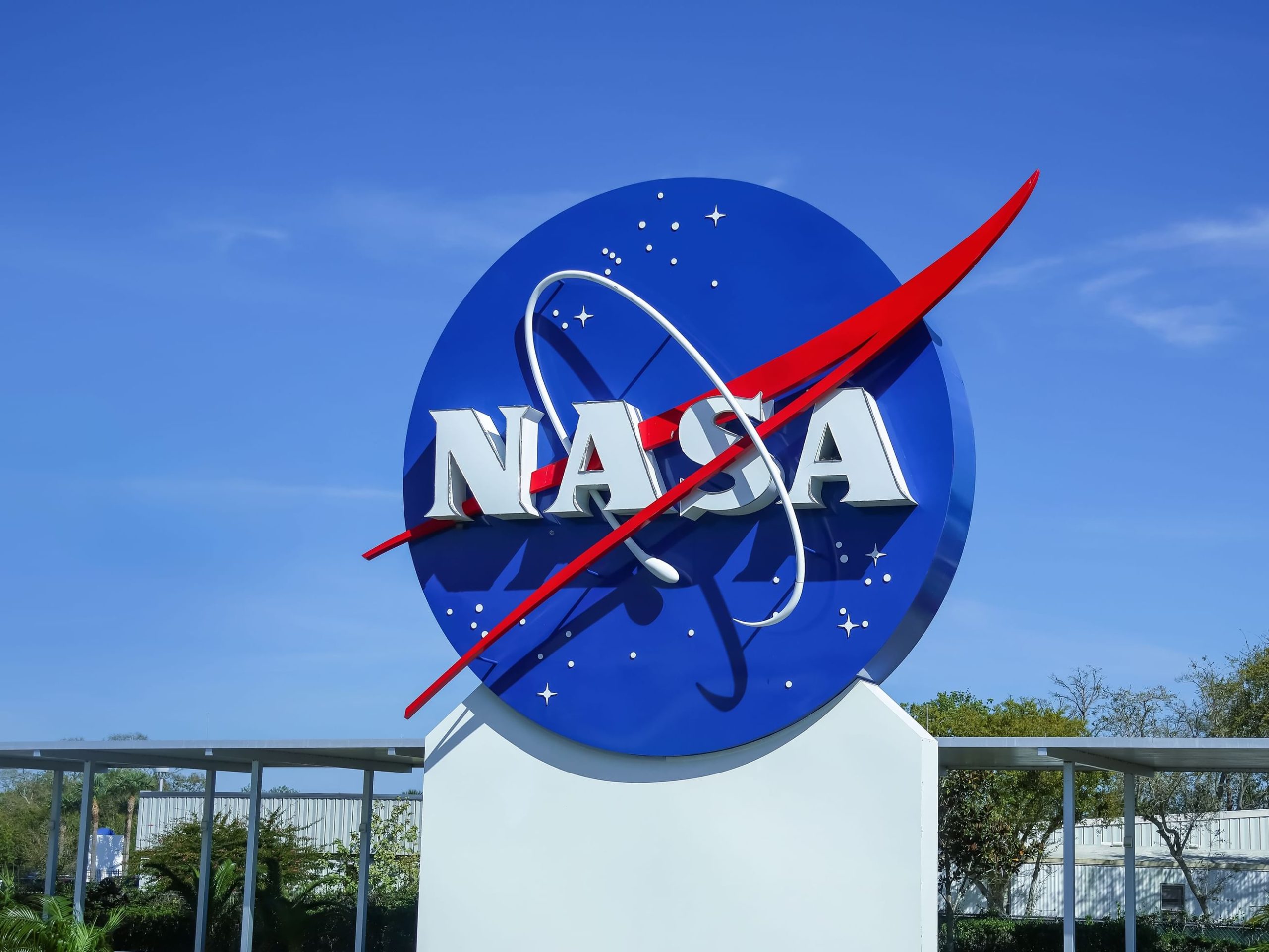 NASA logo sign against a clear blue sky, featuring a blue circle, red chevron, and white letters spelling NASA at the bottom of the emblem.