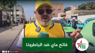 Reporter in a yellow vest and cap speaks into a green mic on a city street; Arabic banner on screen and cars in the background.