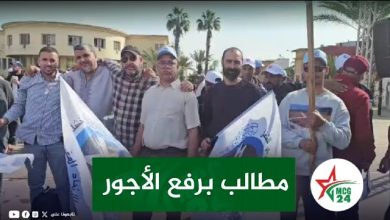 Group of men standing together in a street protest, holding flags, with a green banner that reads 'مطالب برفع الأجور' (Demands to raise wages).