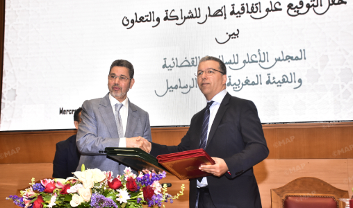 Two men in suits shake hands on a stage, each holding a folder, with a floral arrangement in front and Arabic text on a screen behind them.
