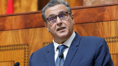 Man in a blue suit and tie with glasses speaks into a microphone in a wood-paneled chamber.