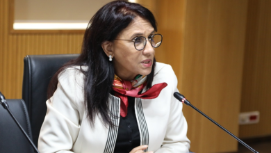 Woman in a white blazer and red scarf speaks into a microphone at a panel discussion in a formal meeting room.