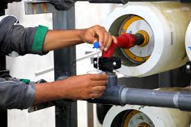 Worker turning a valve handle on a metal pipeline in an industrial setting, with a wrench in hand and colored circular valve wheels nearby.