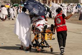 Volunteer in a red vest pushes a patient on a wheeled stretcher shaded by an umbrella at a crowded outdoor event.