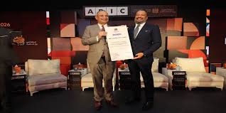 photo | MCG24 | موقع إلكتروني مغربي شامل متجدد على مدار الساعة Two men in suits on a stage exchange a framed certificate during an award ceremony, with a patterned backdrop and seating on stage behind them.