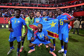 Group of soccer players in blue jerseys with yellow stars celebrate on the field, some kneeling, after a match.