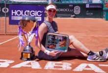 Female tennis player in a gray outfit sits on clay court with a large silver trophy beside her, holding a framed award tablet.
