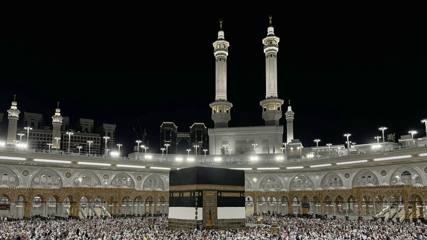 Night view of the Kaaba surrounded by pilgrims at the Grand Mosque (Masjid al-Haram) with illuminated arches and minarets.