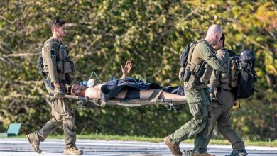 Four soldiers in tactical gear transport a wounded person on a stretcher across a pavement area, outdoors on a sunny day.