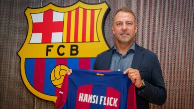 A man in a blue blazer holds up a blue-and-red FC Barcelona jersey in front of a large FC Barcelona shield.