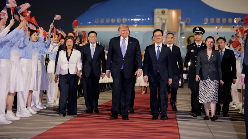 Delegation arrives on a red carpet at an airport, led by a central group of leaders with flag‑bearing attendants nearby.