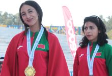 Two female athletes stand on a podium wearing red capes and green uniforms, medals around their necks after a competition, outdoor stadium backdrop.