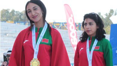 Two female athletes stand on a podium wearing red capes and green uniforms, medals around their necks after a competition, outdoor stadium backdrop.