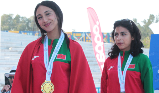 Two female athletes stand on a podium wearing red capes and green uniforms, medals around their necks after a competition, outdoor stadium backdrop.