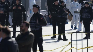 Armed police in black tactical gear stand behind yellow caution tape, with medical personnel in white suits in the background.