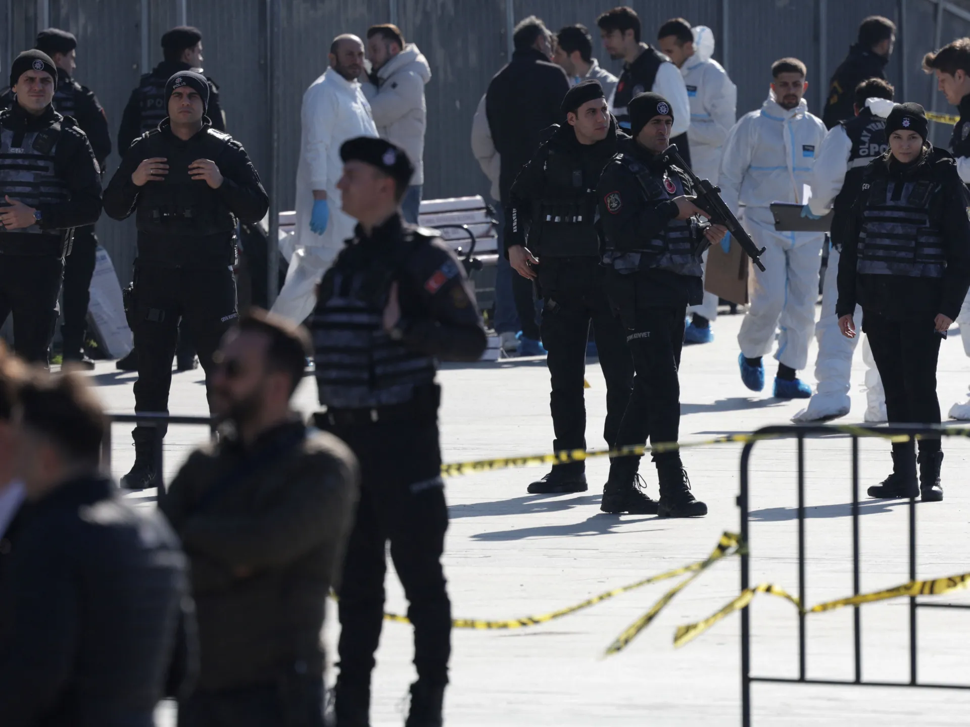 Armed police in black tactical gear stand behind yellow caution tape, with medical personnel in white suits in the background.