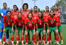 u17jpg | MCG24 | موقع إلكتروني مغربي شامل متجدد على مدار الساعة Senior girls' soccer team posing for a group photo on a sunny field, wearing red jerseys with green shorts and matching socks, numbers visible on their chests.