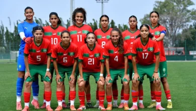 Senior girls' soccer team posing for a group photo on a sunny field, wearing red jerseys with green shorts and matching socks, numbers visible on their chests.