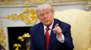 Man in a dark suit and red tie sits in a gilded, ornate room, gesturing with his right hand and pointing.