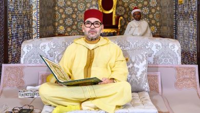 Man in a yellow traditional robe and red fez sits cross‑legged on a patterned sofa, reading a large green book; a second person in white sits in the background.