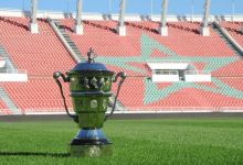 Polished trophy on a green soccer field with red stadium seats and a large turquoise star design in the background.
