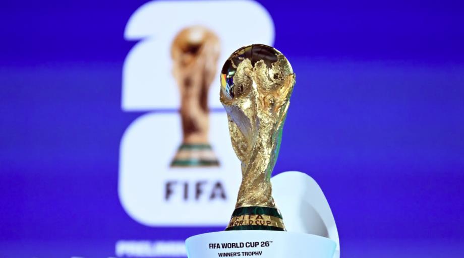 Golden FIFA World Cup trophy on a white pedestal against a blue backdrop, foreground in sharp focus.