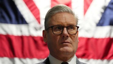 Man in a suit and glasses standing in front of a large Union Jack flag ground behind him?