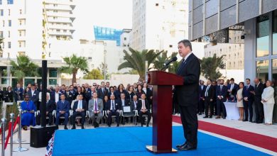 Man in a suit speaks at a podium on a blue-carpeted outdoor stage as a formal ceremony unfolds, with a crowd behind him.