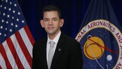 Man in a suit stands before a U.S. flag and a NASA banner, likely at a press event or briefing.