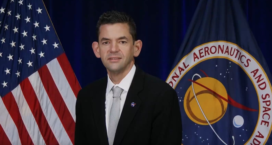 Man in a suit stands before a U.S. flag and a NASA banner, likely at a press event or briefing.