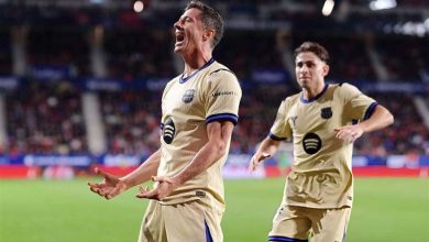 Two soccer players in beige uniforms celebrate on a green field in a stadium, one shouting with arms outstretched in triumph.