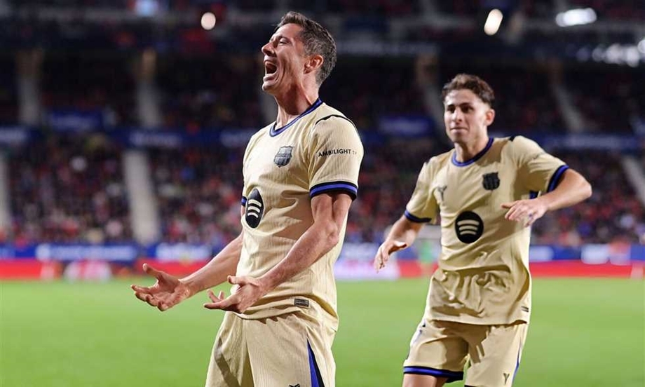 Two soccer players in beige uniforms celebrate on a green field in a stadium, one shouting with arms outstretched in triumph.