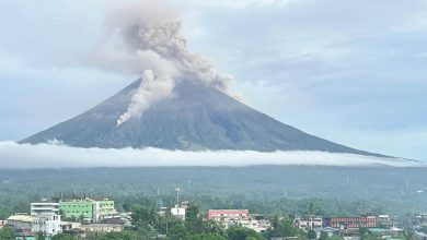 Active volcano erupting with a tall ash plume, looming over a town and green valley below.