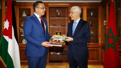 Two men in suits exchange a magazine in a formal office, with Jordan and Morocco flags visible in the background.