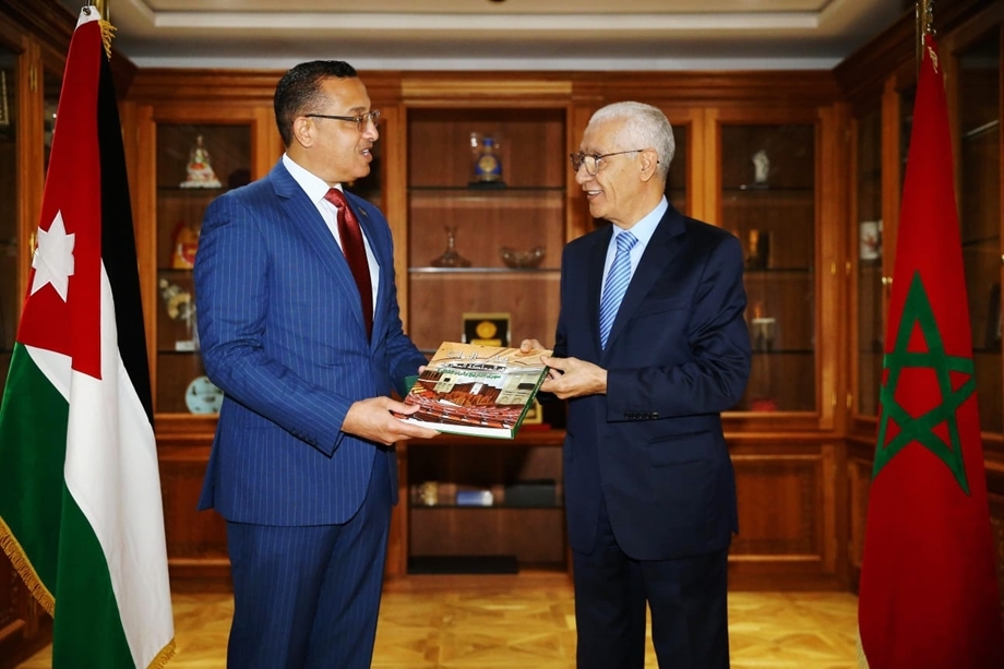 Two men in suits exchange a magazine in a formal office, with Jordan and Morocco flags visible in the background.