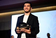 Man on stage in a dark blazer and white shirt, smiling while holding a trophy award toward the audience on a well-lit stage