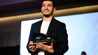 Man on stage in a dark blazer and white shirt, smiling while holding a trophy award toward the audience on a well-lit stage