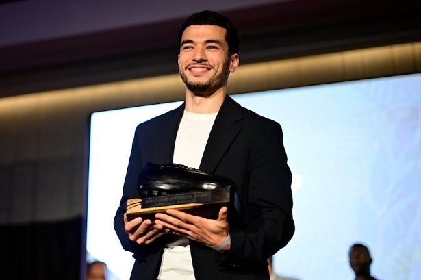 Man on stage in a dark blazer and white shirt, smiling while holding a trophy award toward the audience on a well-lit stage