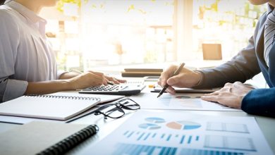 Two professionals analyze financial reports and charts at a sunlit desk, with a calculator, notebook, and glasses nearby.