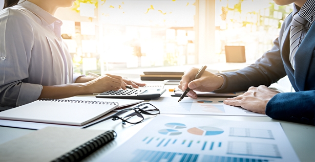 Two professionals analyze financial reports and charts at a sunlit desk, with a calculator, notebook, and glasses nearby.