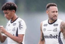 Two male soccer players in Santos FC training shirts on a practice field; left player checks a phone, right looks ahead.