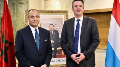Two men in dark suits pose for a formal photo in an office, with a framed portrait and flags on either side behind them.