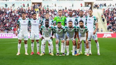 Soccer team in white jerseys poses for a group photo on the field, with fans in the stands behind them.