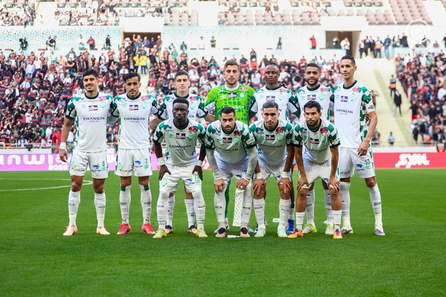 Soccer team in white jerseys poses for a group photo on the field, with fans in the stands behind them.