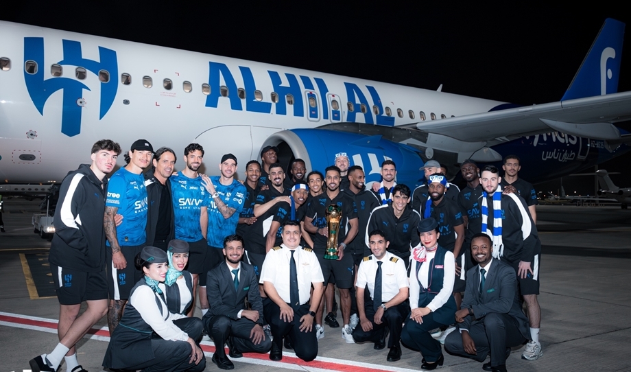 Group of airline crew and athletes posing with a trophy beside a blue-and-white airplane at night on the tarmac.