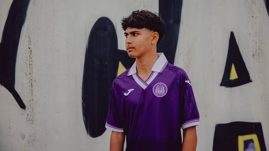 Young man in a purple soccer jersey stands in front of a graffiti-covered wall.