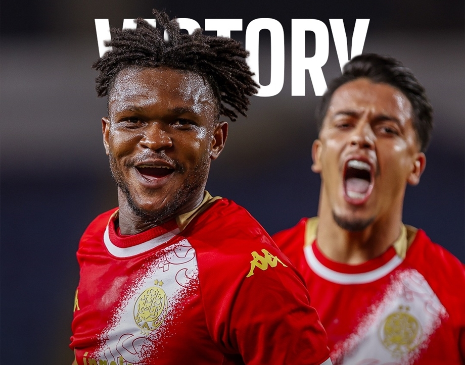 Two footballers in red kits celebrate a victory; the front player smiles at the camera while another teammate shouts in the background, with 'VICTORY' visible above.