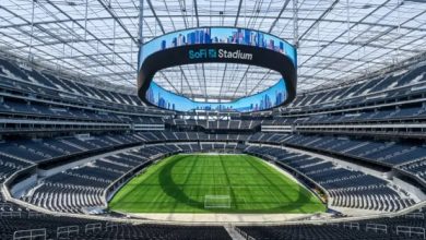 Inside SoFi Stadium, a circular video board hangs above a bright green field with empty dark seats surrounding it.