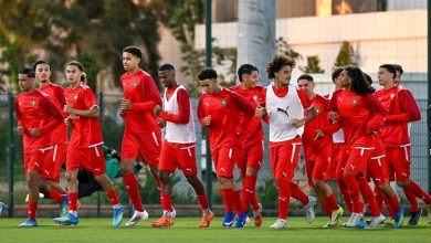 A group of young male soccer players in red uniforms jog together on a green training field during practice.