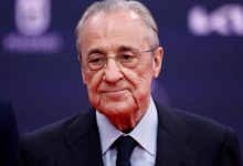 Older man in a dark suit and tie, wearing glasses, looking slightly downward with a neutral expression at a formal event.